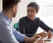 Young indian female mentor coach worker talking to male coworker teaching intern having business conversation with workmate, serious hindu manager helping colleague discussing new project in office