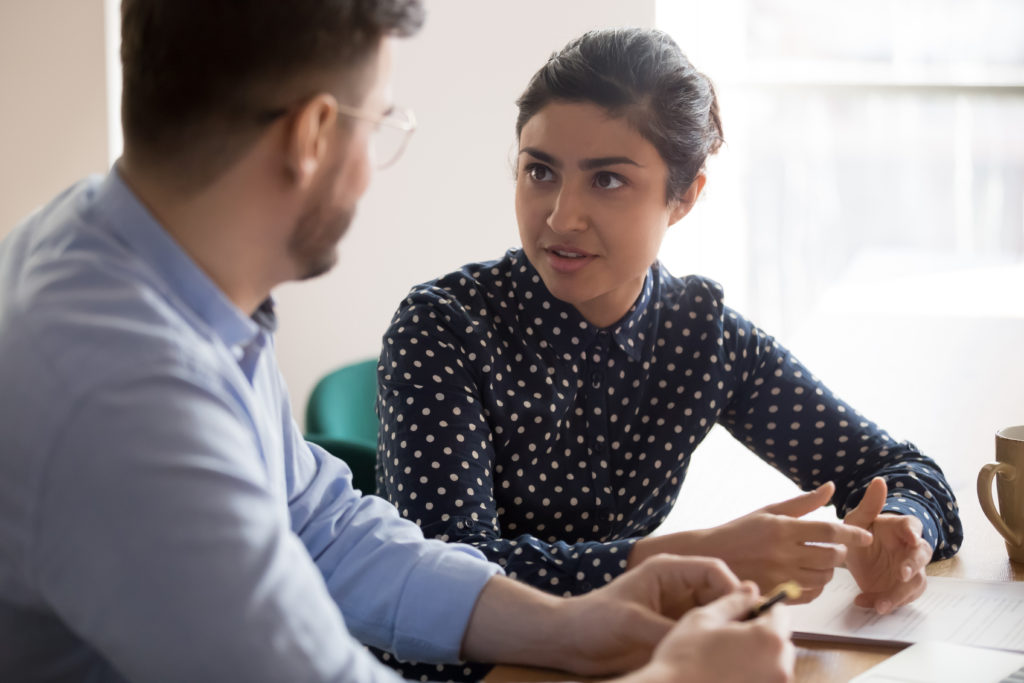 Young indian female mentor coach worker talking to male coworker teaching intern having business conversation with workmate, serious hindu manager helping colleague discussing new project in office